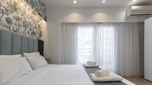 A modern hotel room with a large bed, a headboard with a floral pattern, a window with sheer curtains, and an air conditioning unit on the wall. - Kriel Suites by LIV Homes 202 (Athens)