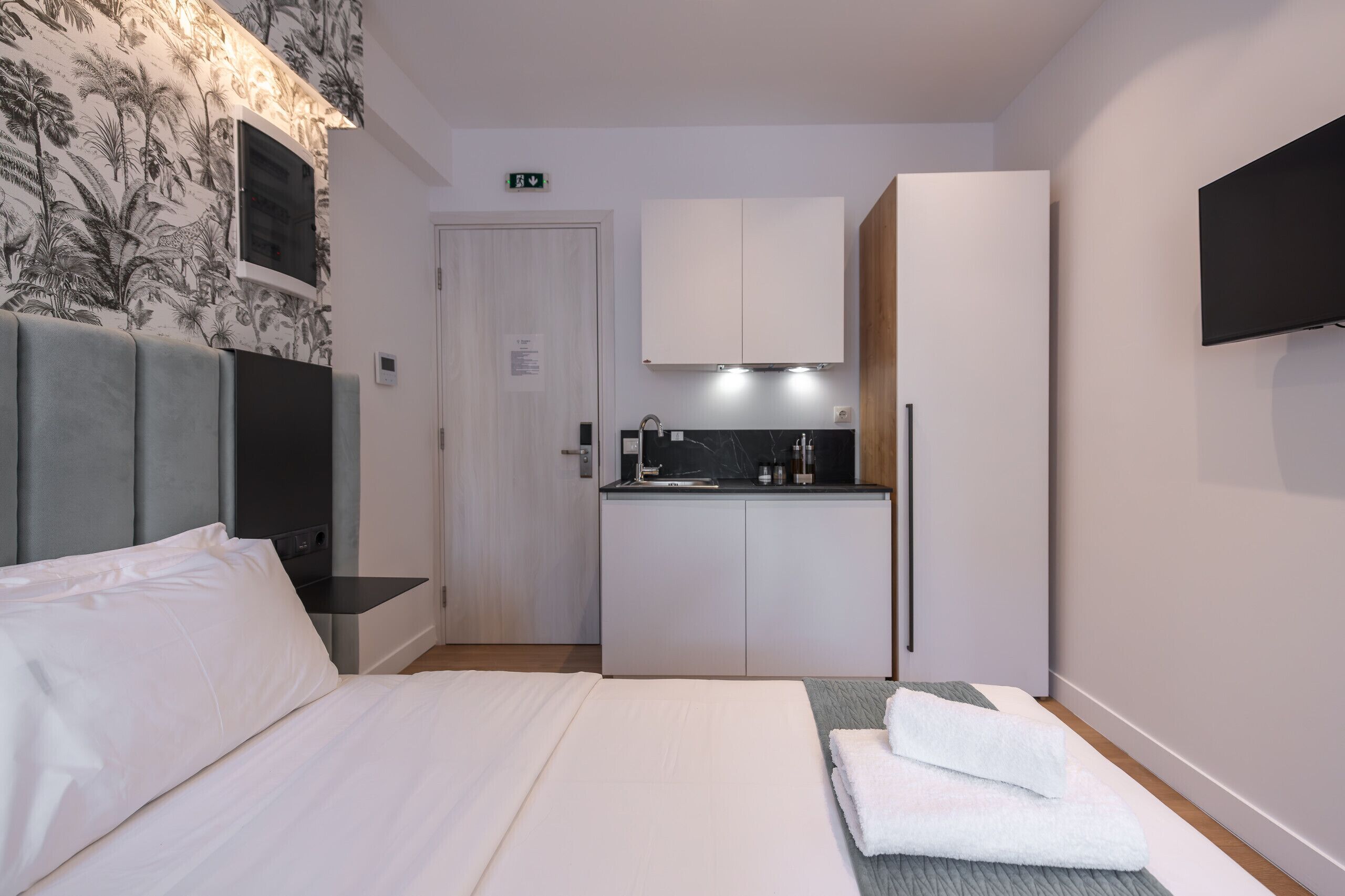 A compact hotel room with a bed, a small kitchenette, a TV mounted on the wall, and a door with an emergency exit sign.