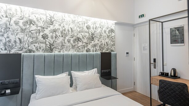 A modern hotel room with a large bed, a headboard with built-in controls, a mirror, a small table with a kettle, and a wooden ceiling. - Kriel Suites by LIV Homes 304 (Athens)