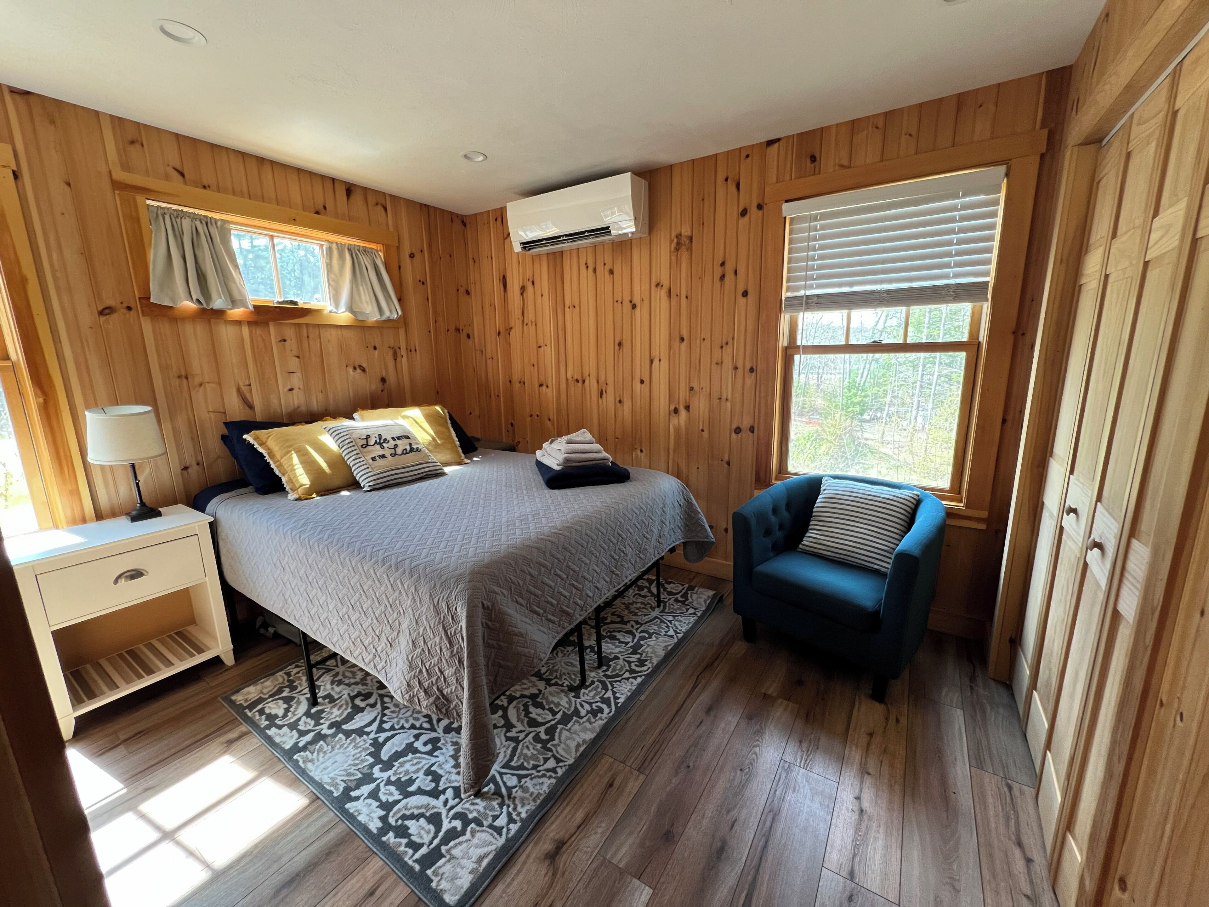 A wooden-paneled bedroom with a bed, bedside table, chair, and large windows.