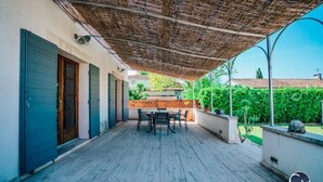 A covered patio with a table and chairs, a thatched roof, and a view of a garden and buildings in the background. - Maison Avec Piscine, Jacuzzi, Sauna et Billard (Le Thor)