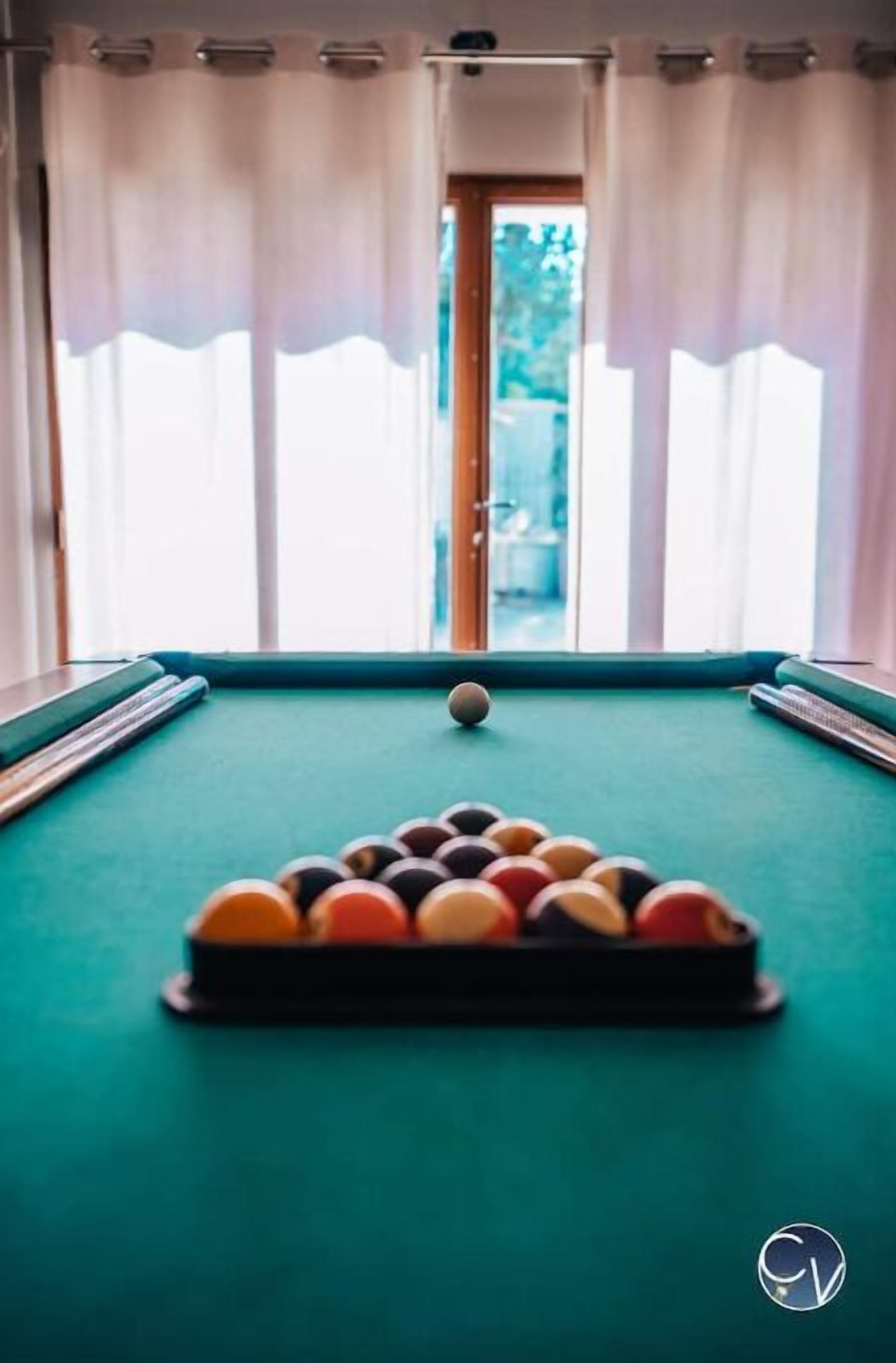 Une table de billard avec les billes disposées en forme de triangle, un queue de billard et une fenêtre avec des rideaux.