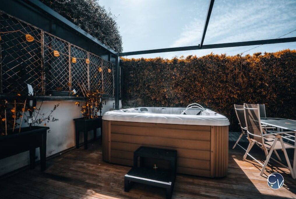 Un jacuzzi en un espacio exterior cubierto con suelo de madera, una mesa y sillas, y un muro de seto.