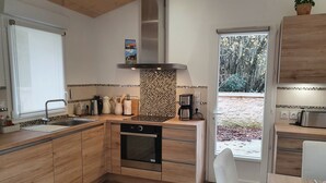 A modern kitchen with wooden cabinets, a built-in oven, a coffee maker, and a view of the garden through the window. - Petit Paradis Avec Piscine Partagée (Gizay)