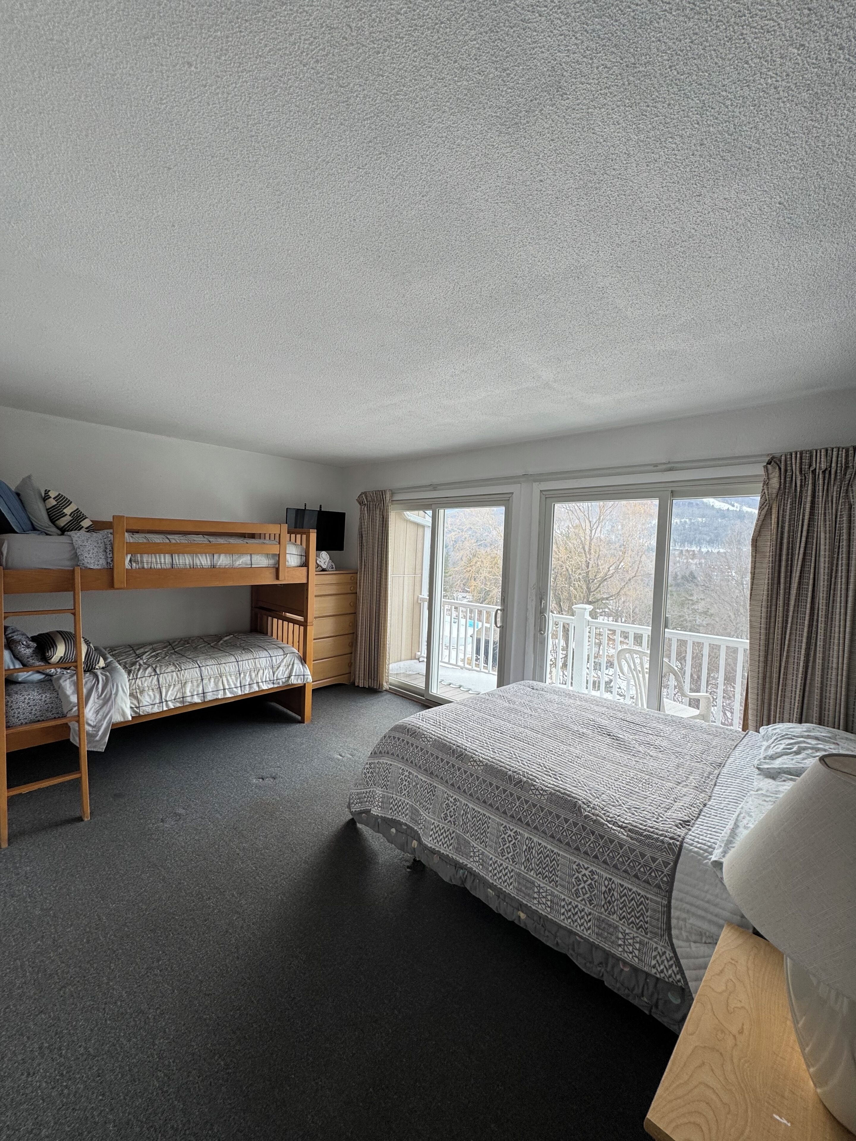 Une chambre avec un lit superposé, un lit simple, un bureau et une fenêtre donnant sur un balcon.