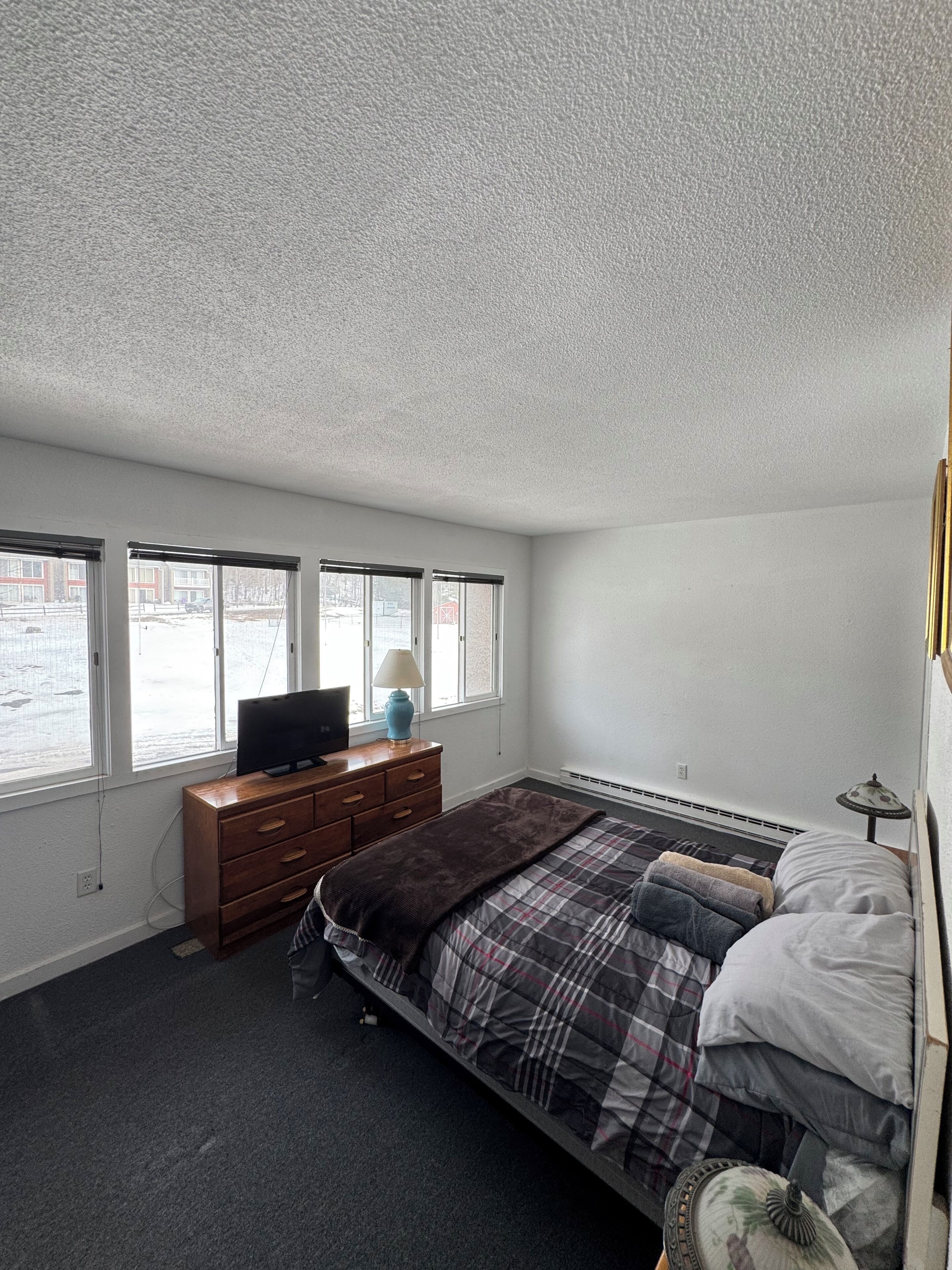 Une chambre à coucher comprenant un lit, une commode, un téléviseur et une fenêtre donnant sur des immeubles.