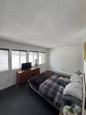 Une chambre à coucher comprenant un lit, une commode, un téléviseur et une fenêtre donnant sur des immeubles.