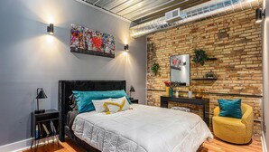 Un dormitorio moderno con pared de ladrillo, una cama grande con ropa blanca, un sillón amarillo y un cuadro en la pared.