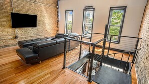 Una sala de estar moderna con un sofá de cuero negro en forma de L, un televisor de pantalla plana montado en una pared de ladrillo y una escalera con barandilla de vidrio que lleva a un nivel superior.