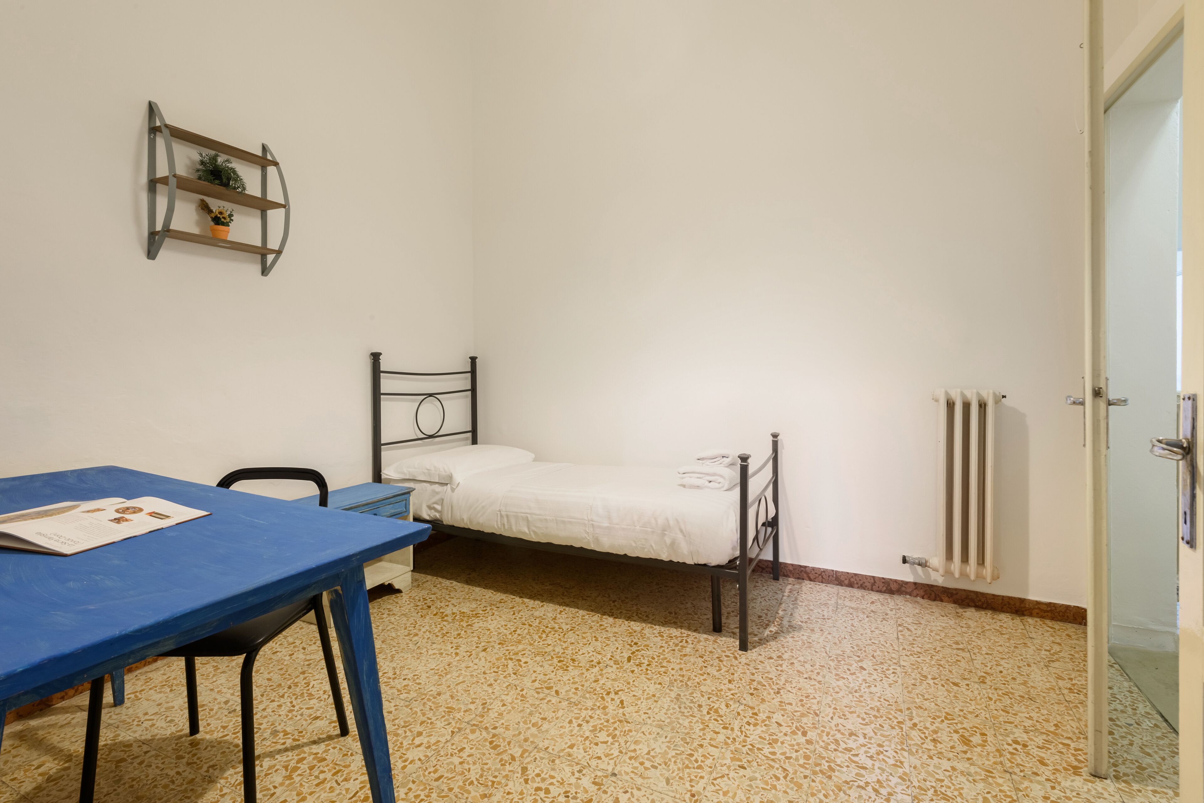 Ein schlichter, einfach eingerichteter Raum mit einem Bett, einem Tisch und einem Stuhl. An der Wand ist ein Heizkörper zu sehen, oberhalb des Bettes ist ein Regal mit einer Pflanze angebracht.