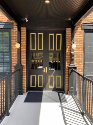 A black double door with gold accents and glass panels, leading to a brick building with a black roof and a metal railing. - Ulysses Suites, Suite 201, 2 bedroom/2 bath, full kitchen and living room (Galena)