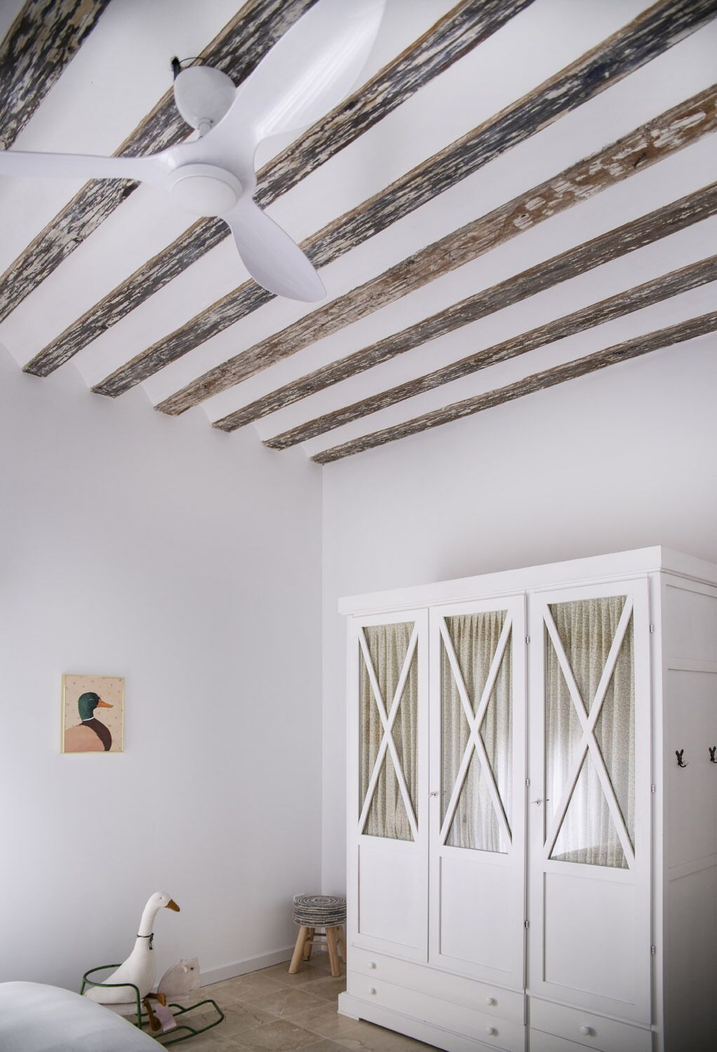 A room with a white ceiling fan, exposed wooden beams, a white wardrobe with glass doors, a duck-shaped chair, and a small stool.