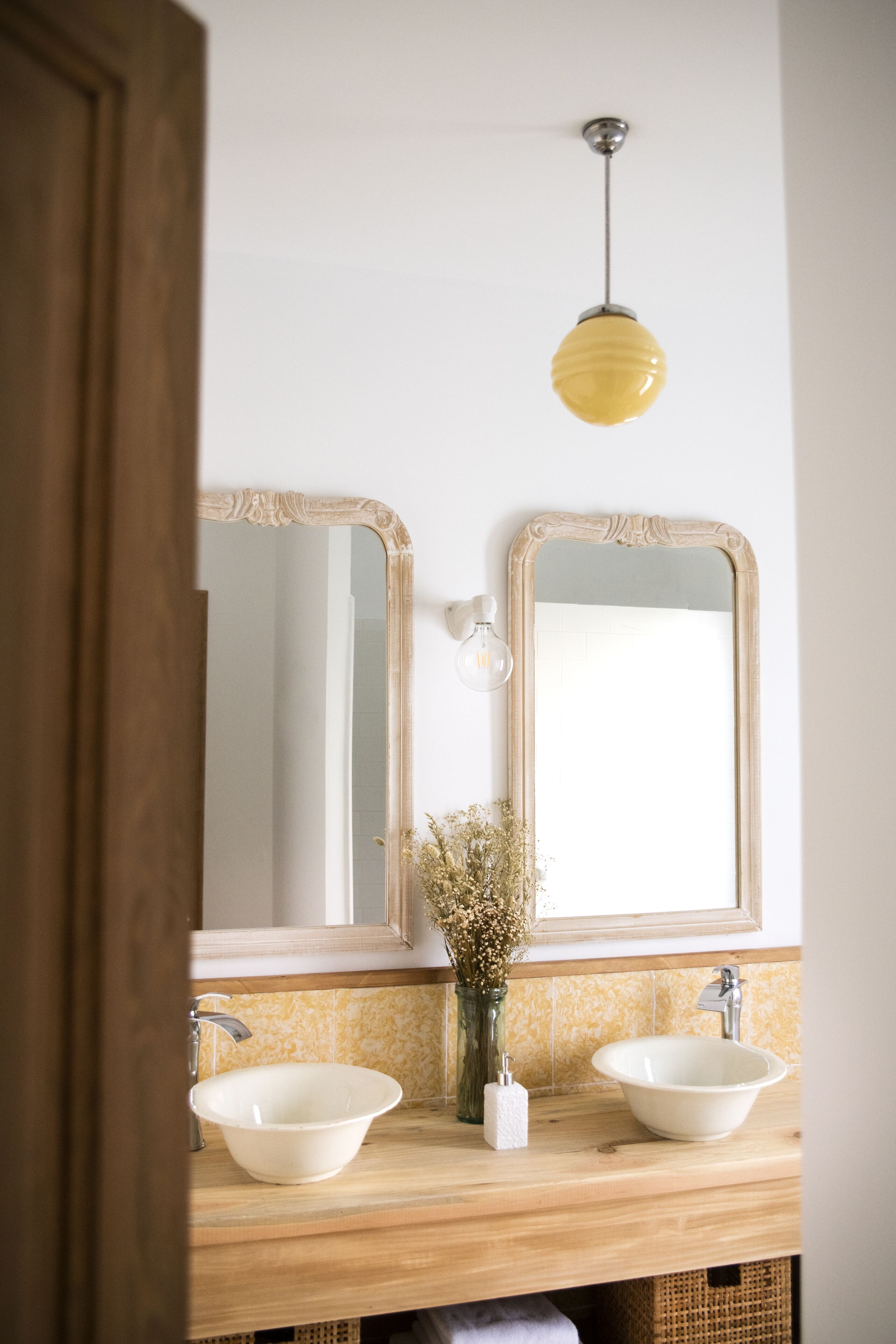 Een badkamer met twee wasbakken, een spiegel, een lamp en een vaas met gedroogde bloemen.