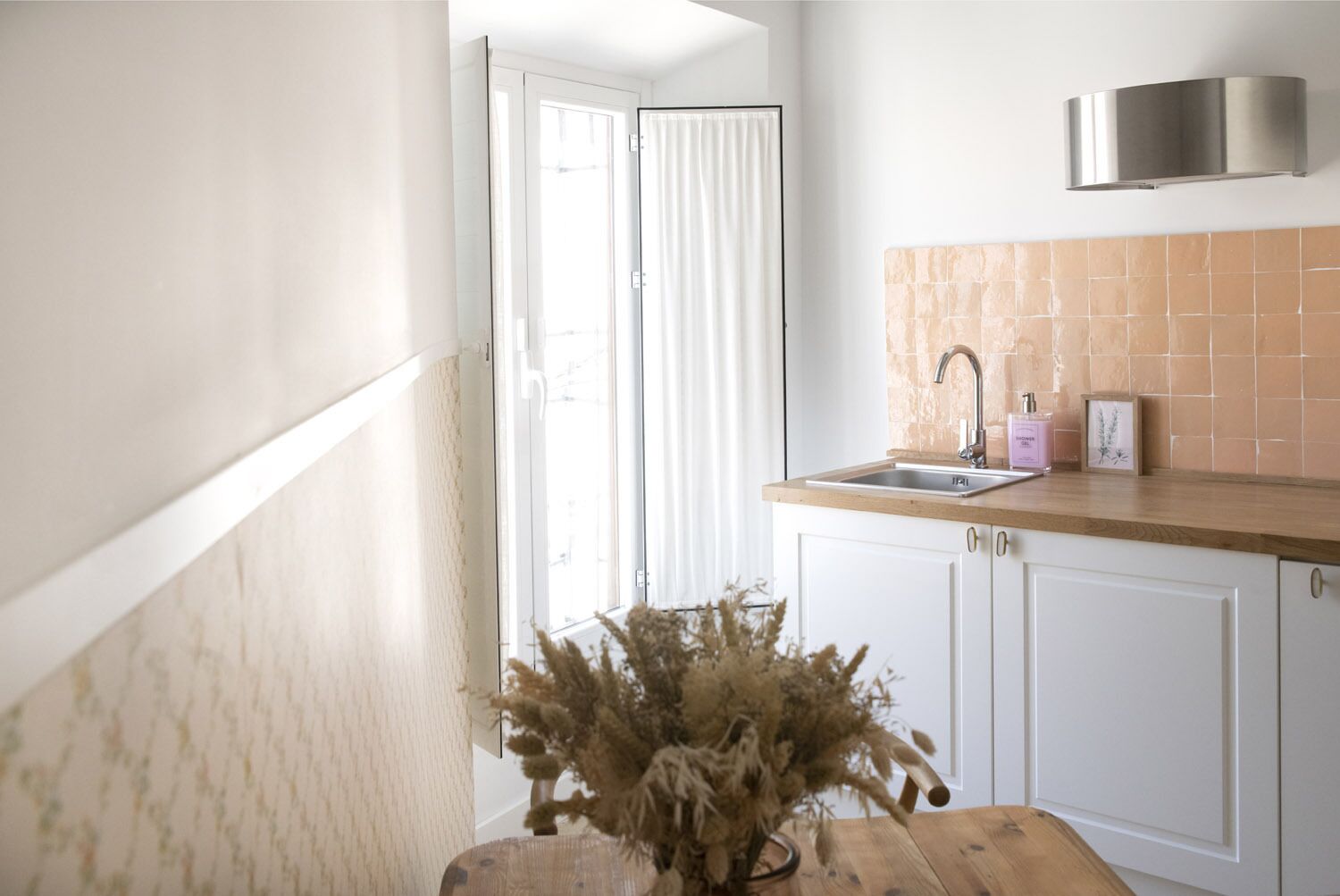 Una cocina con armarios blancos, encimera de madera, pileta de acero inoxidable y un jarrón decorativo con plantas secas.