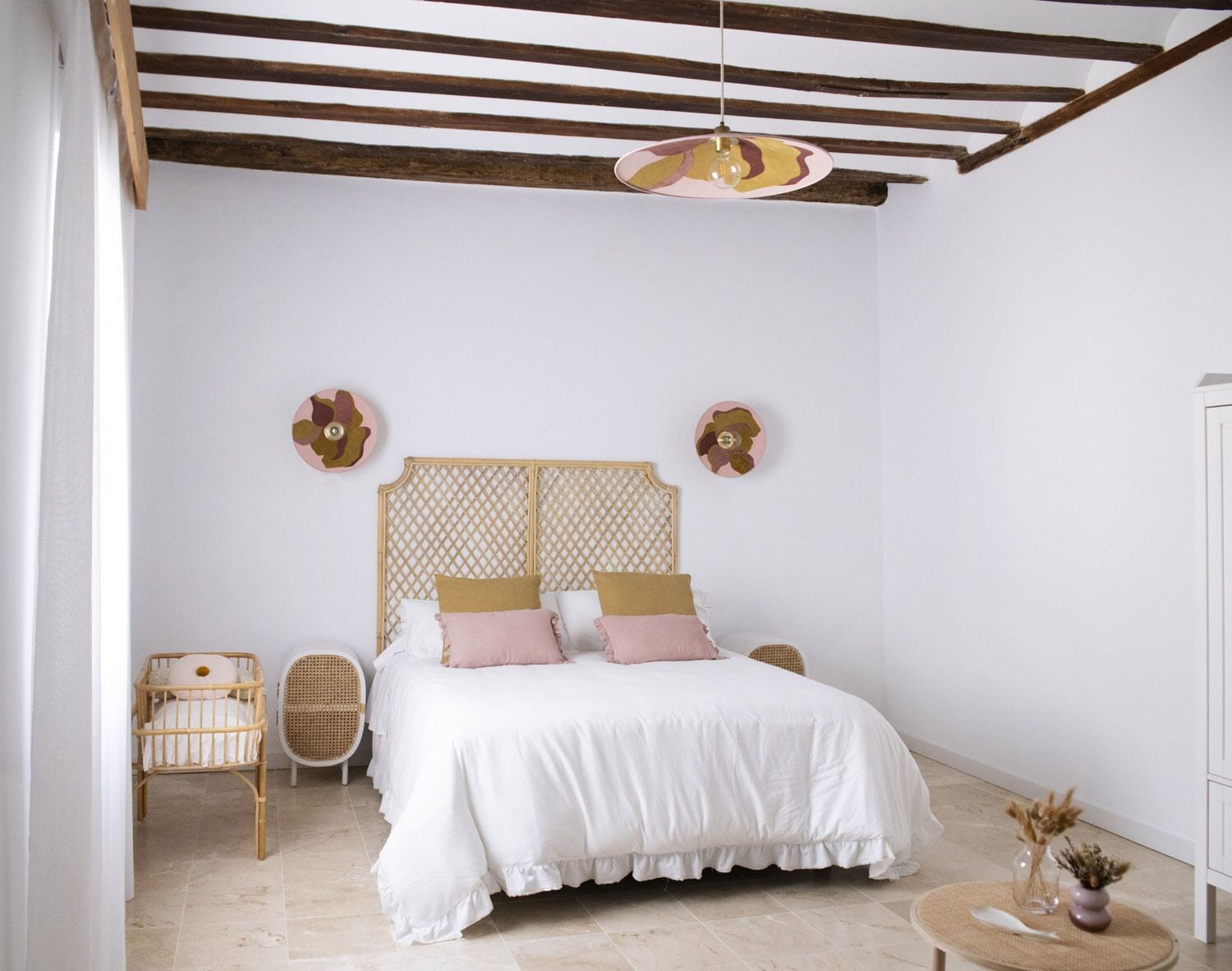 Un dormitorio con una cama, dos sillas de mimbre, una mesa redonda con un jarrón y una lámpara colgante.