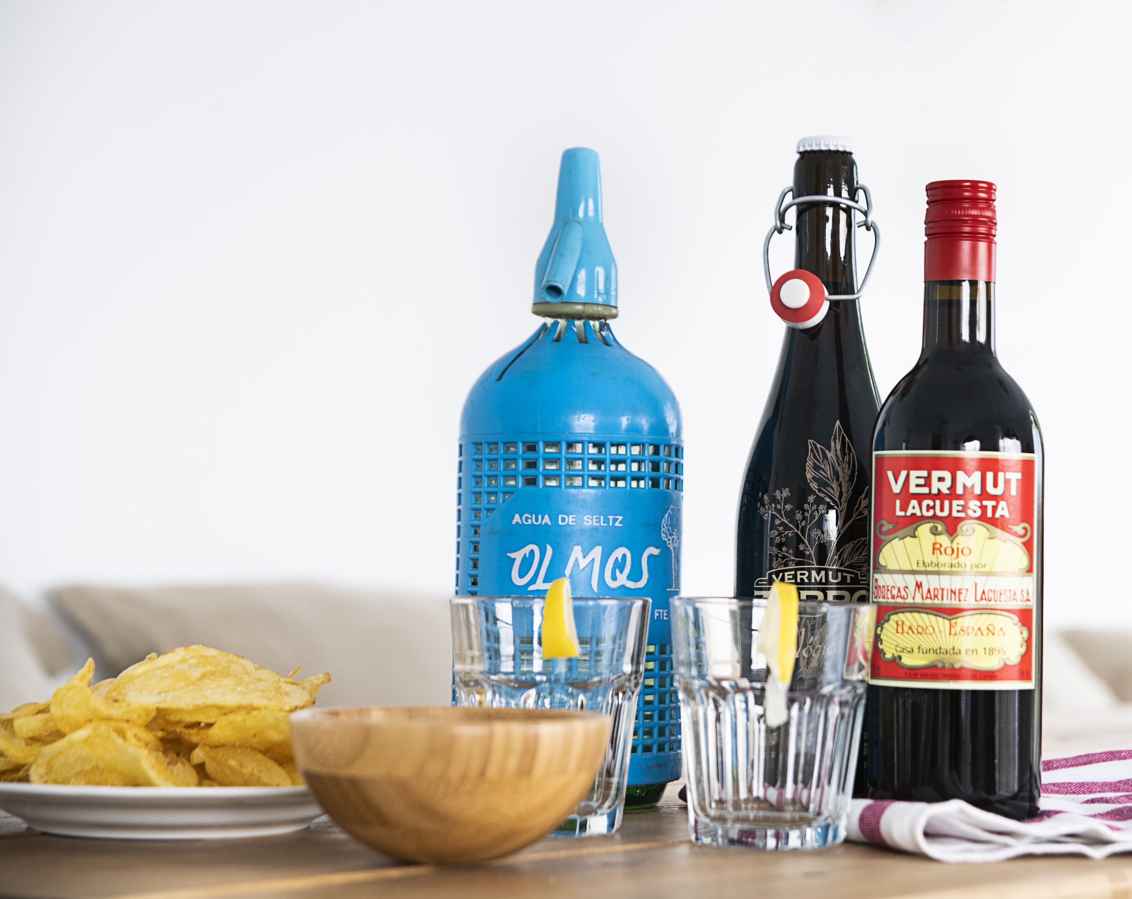 Een tafel met een bord chips, een houten schaal, een blauwe waterfles, een glas met een citroenpartje, een fles Vermut Lacusta en een fles rode wijn.