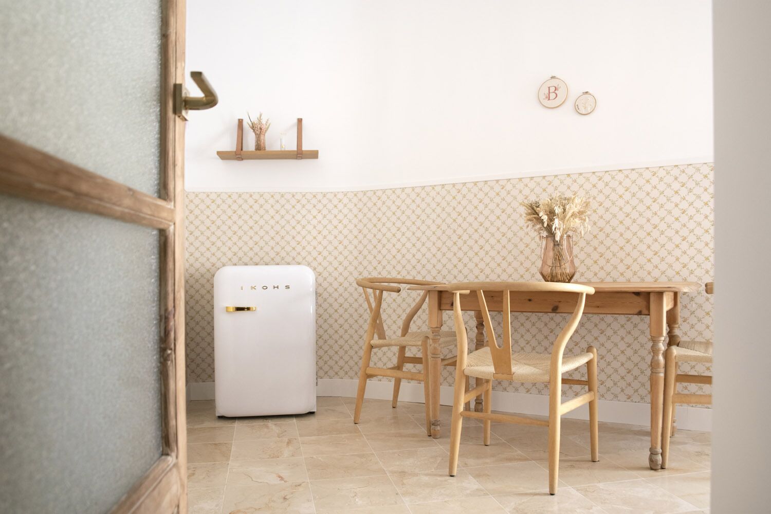 Una cocina con refrigerador blanco, mesa de comedor de madera y sillas. Hay una estantería con adornos y una pared con papel tapiz estampado.