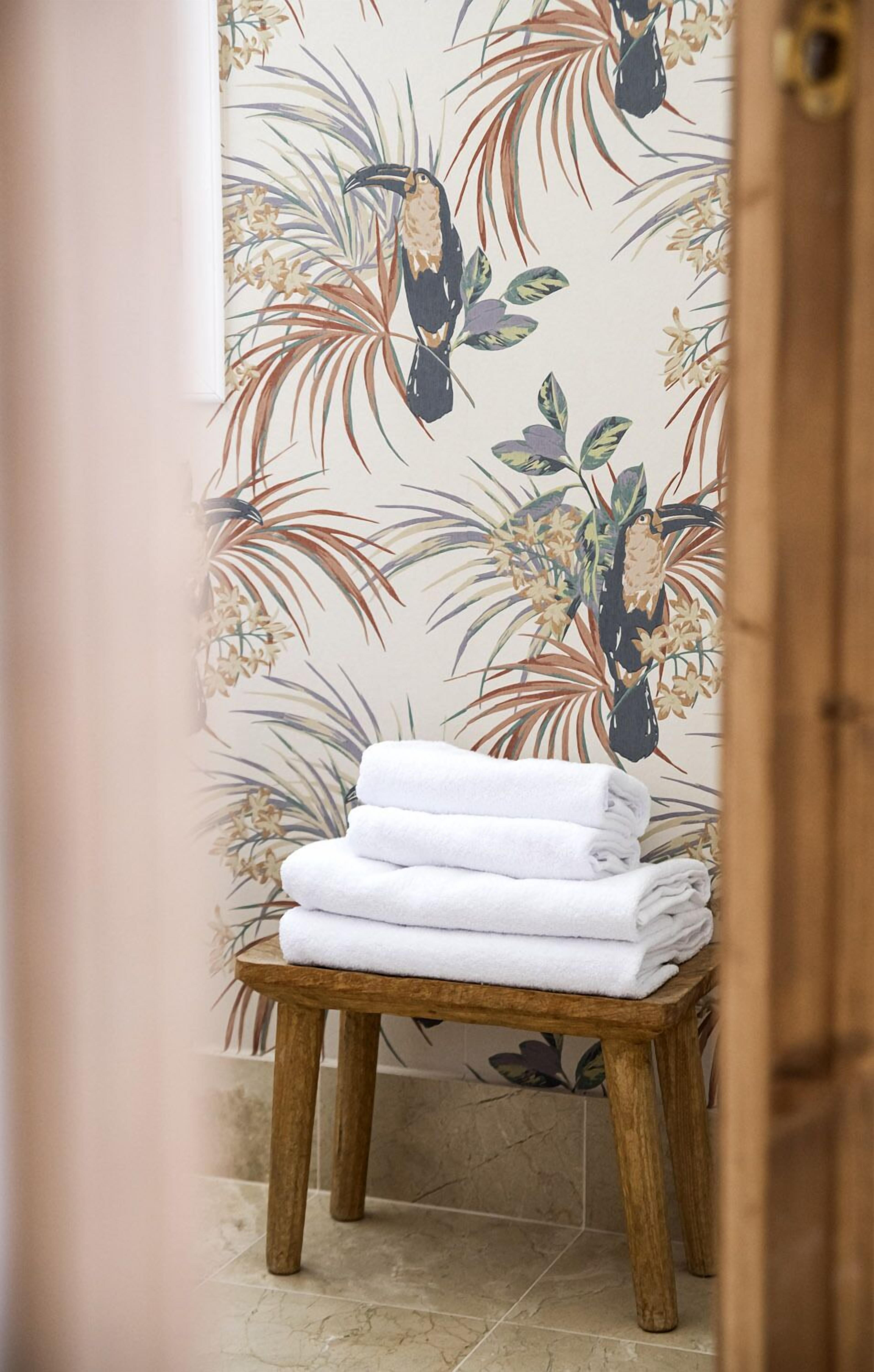 A bathroom with a wooden stool holding white towels, a floral wallpaper, and a tiled floor.