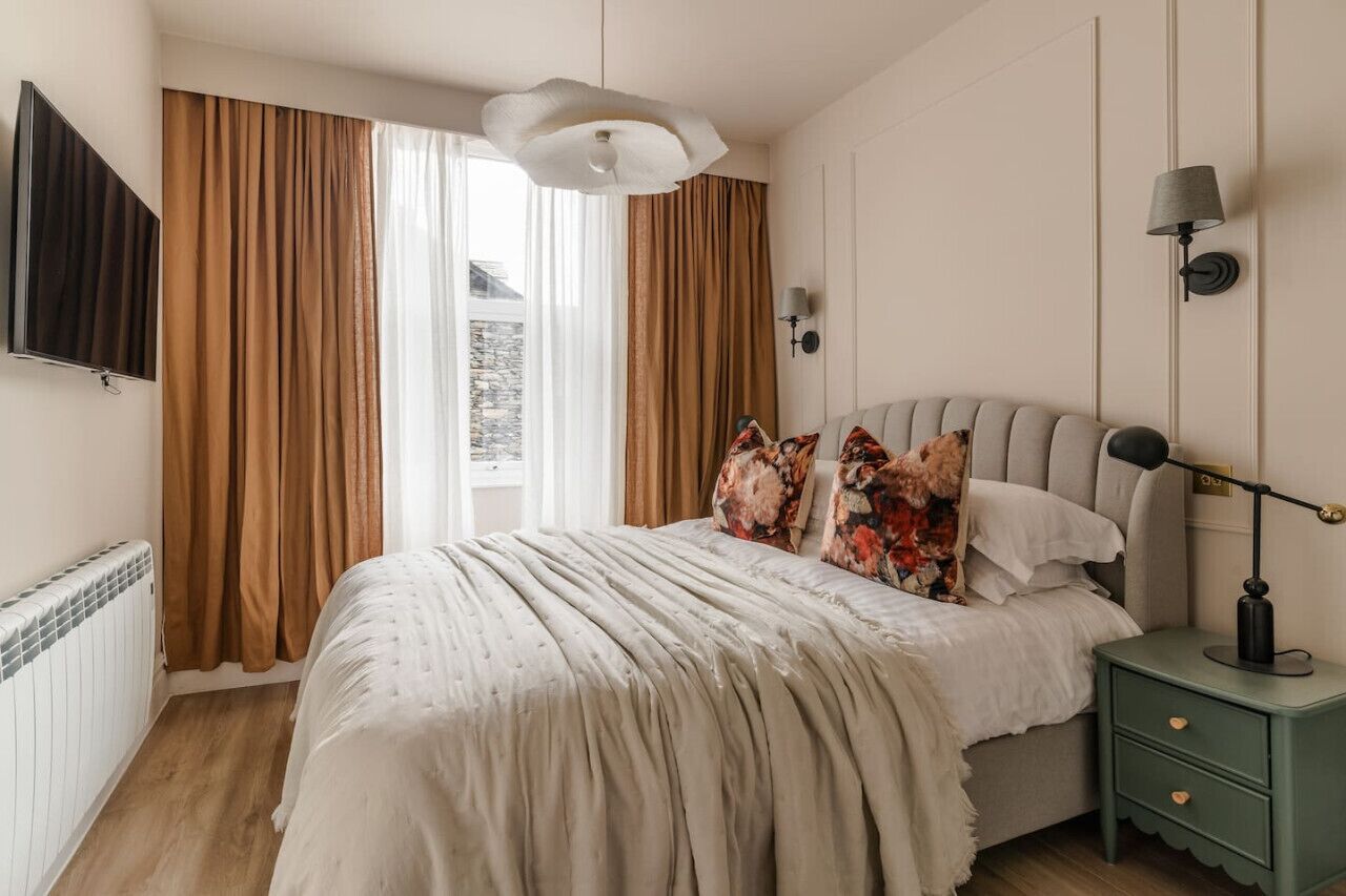 Une chambre à coucher avec un lit, des rideaux, une lampe, un téléviseur et une vue sur l’extérieur.