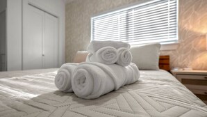 A neatly made bed with rolled white towels and a pillow, a bedside table with a lamp, and a window with blinds. - Island Gems - Gold

 (Galveston)