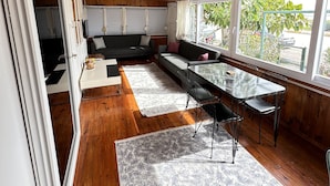 A modern interior with a wooden ceiling, a sofa, a dining table with chairs, and large windows offering a view of greenery outside.
