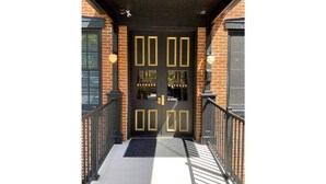 A black double door with gold accents and glass panels, leading to a brick building with a black roof and a metal railing. - Ulysses Suites, Suite 203 (Galena)