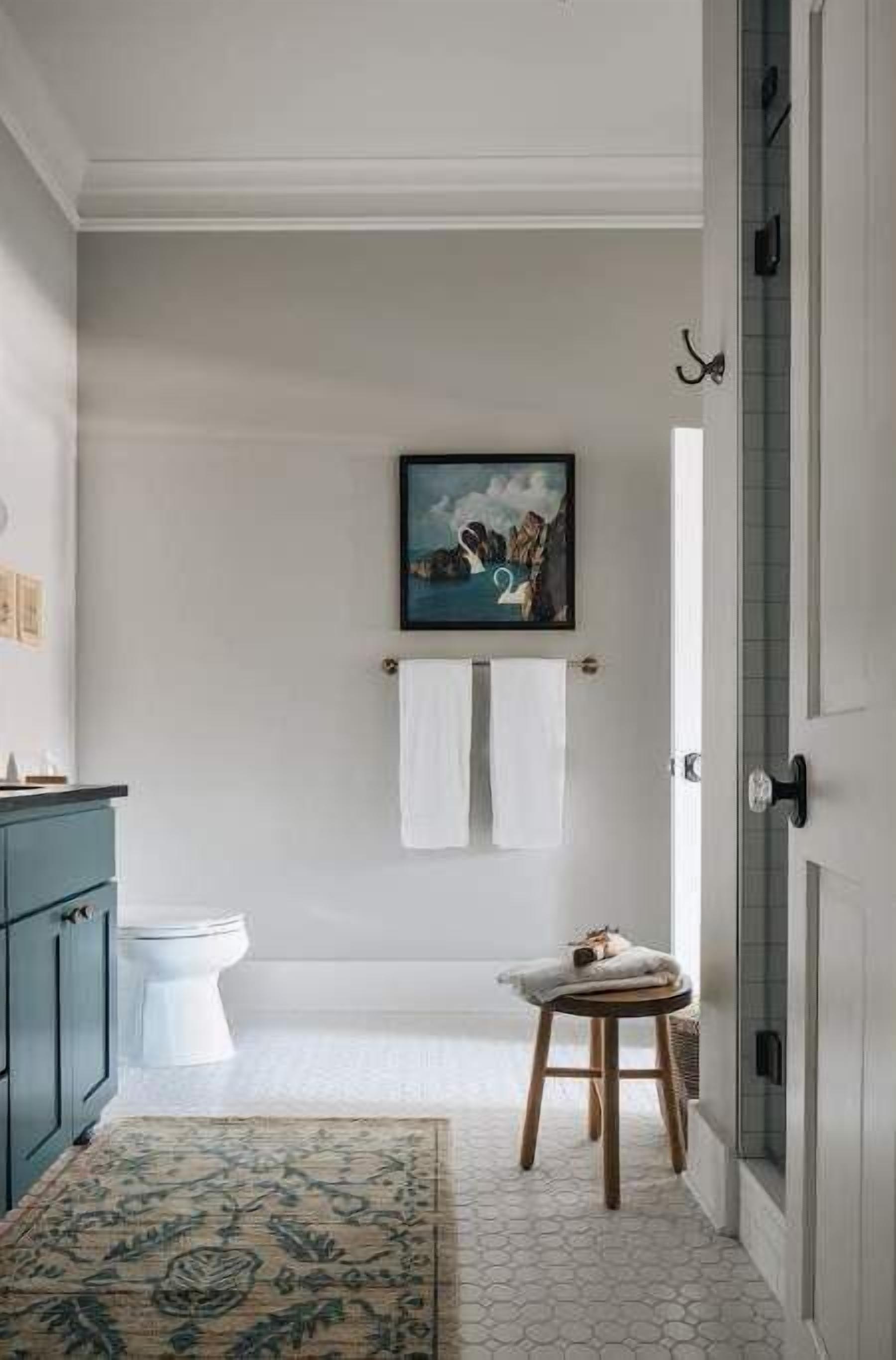 A bathroom with a toilet, a sink, a mirror, a towel rack with two white towels, a framed picture on the wall, and a patterned rug on the floor.