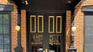 A black double door with gold accents and glass panels, leading to a brick building with a black roof and a metal railing.