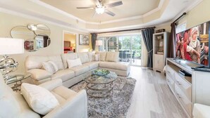 A spacious living room with a large L-shaped sofa, a glass coffee table, a ceiling fan, and a television. - GolfView Condo w/ $50 ThemePark Credit Near Disney (Reunion)