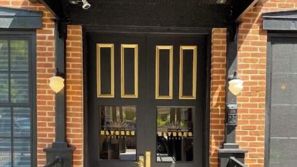 A black double door with gold accents and a glass panel, leading to a brick building with a black roof and a black railing. - Ulysses Suites, Suite 204 (Galena)
