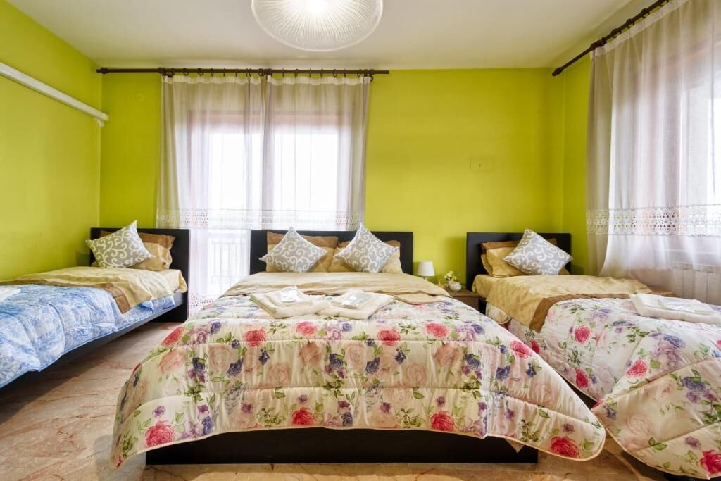 A room with three beds, each with floral bedding, a large window with sheer curtains, and a ceiling fan.