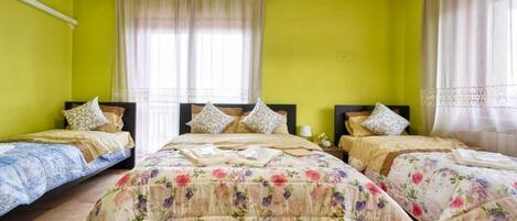A room with three beds, each with floral bedding, a large window with sheer curtains, and a ceiling fan.