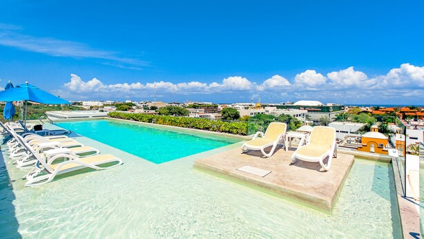 A clear blue pool with lounge chairs and umbrellas, surrounded by buildings and a clear sky. - ANAH - Central Condo - 2 Bedrooms (Playa del Carmen)