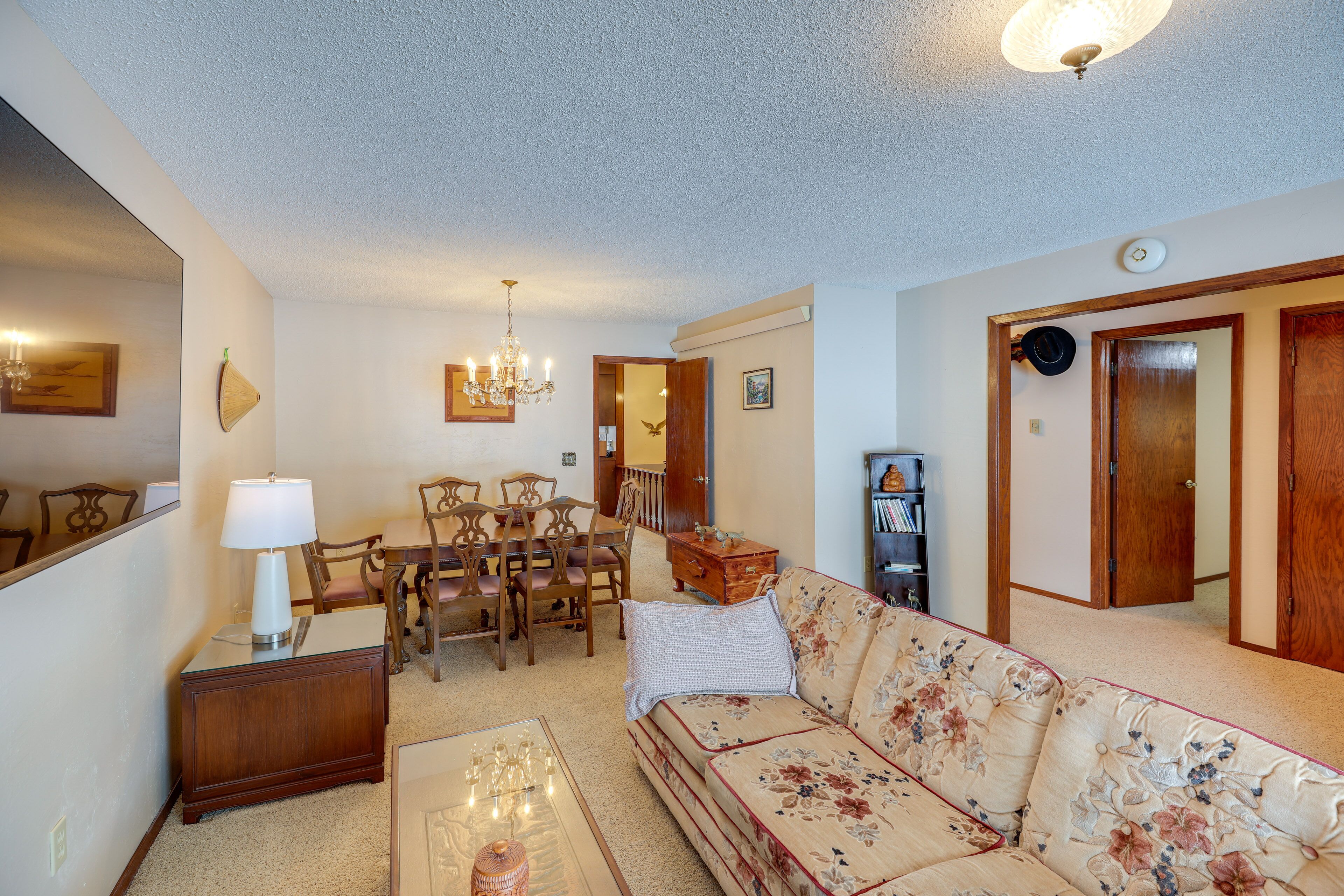 A living room with a floral sofa, wooden dining table, and chairs. There is a chandelier, a lamp, and a bookshelf.