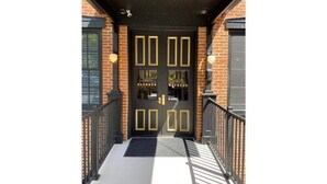 A black double door with gold accents and a glass panel, leading to a brick building with a black roof and a black railing. - Ulysses Suites, Suite 302 (Galena)