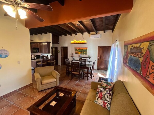 A spacious living area with a sofa, a coffee table, and a dining table set for four. A ceiling fan and a chandelier are visible.