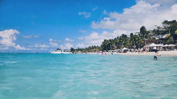 A beach with clear turquoise water, a white boat, and a sandy shore with palm trees and beachgoers. - Sueño Escondido-West Bay Beach (West Bay)