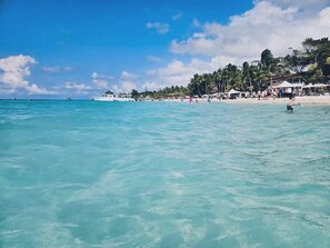 A beach with clear turquoise water, a white boat, and a sandy shore with palm trees and beachgoers.
