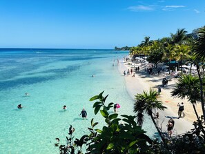 Een strand met helder turquoise water, mensen die zwemmen en over het strand lopen, en palmbomen langs de kust.