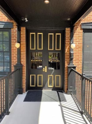 A black double door with gold accents and a glass panel, leading to a brick building with a black roof and a black railing. - Ulysses Suites, Suite 303 (Galena)
