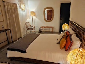 A spacious bedroom with a large bed, a bedside table, a chair, a bench, and a mirror on the wall.