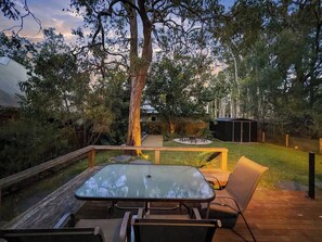 An outdoor deck with a glass-top table and chairs, surrounded by a wooden deck and a grassy area with trees. - Pearl Beach House - 2 min to sand & sea - 2 semis (Pearl Beach)