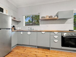 A modern kitchen with grey cabinets, a wooden countertop, a stainless steel refrigerator, a built-in oven, and a window with a view of trees.
