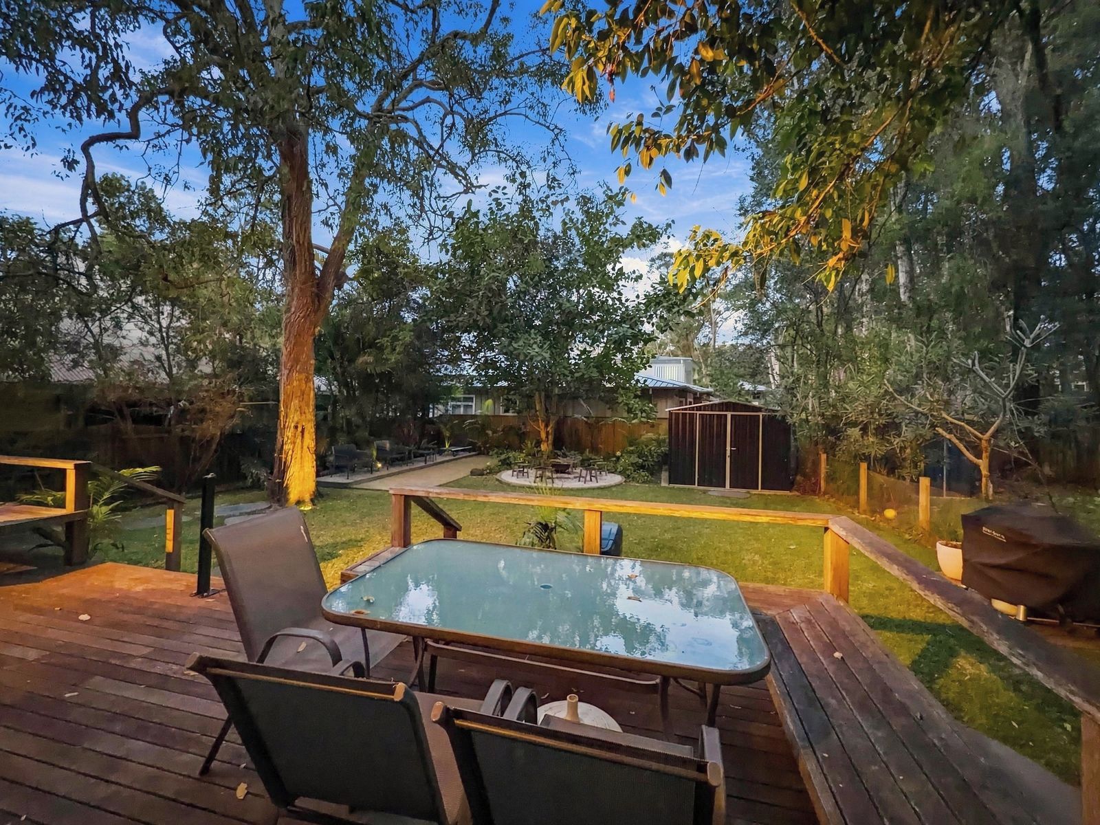 Eine Terrasse im Freien mit einem Tisch und Stühlen mit Glasplatte, umgeben von Bäumen und einer Rasenfläche.
