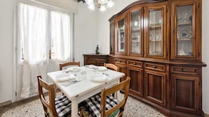 Une salle à manger avec une table blanche dressée pour quatre personnes, entourée de chaises en bois dotées de coussins à carreaux. Un buffet en bois, avec des portes en verre, est adossé au mur.