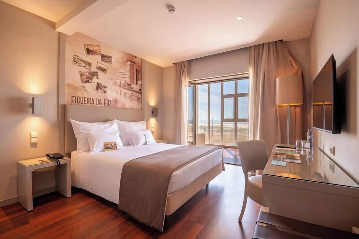 Una habitación de hotel con cama, mesita de noche, escritorio, silla y un ventanal grande.