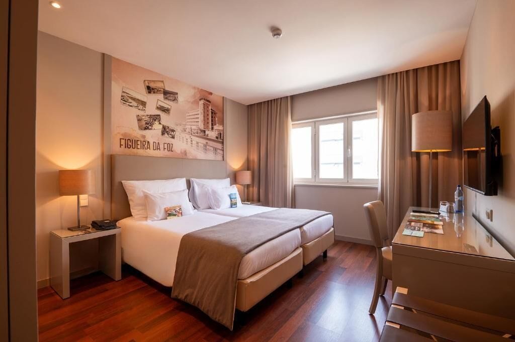Habitación de hotel con cama, mesitas de noche, escritorio, silla, televisión y ventana con cortinas.