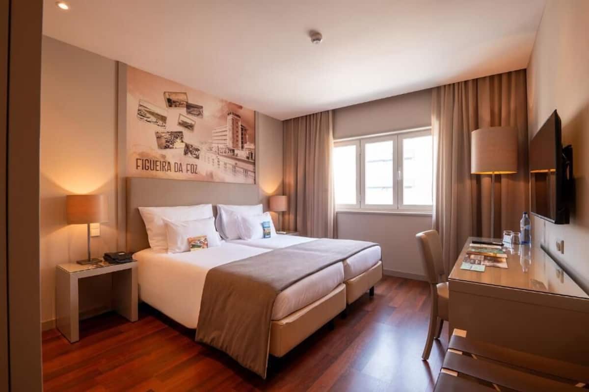 Habitación de hotel con cama, mesitas de noche, escritorio, silla, televisión y ventana con cortinas.