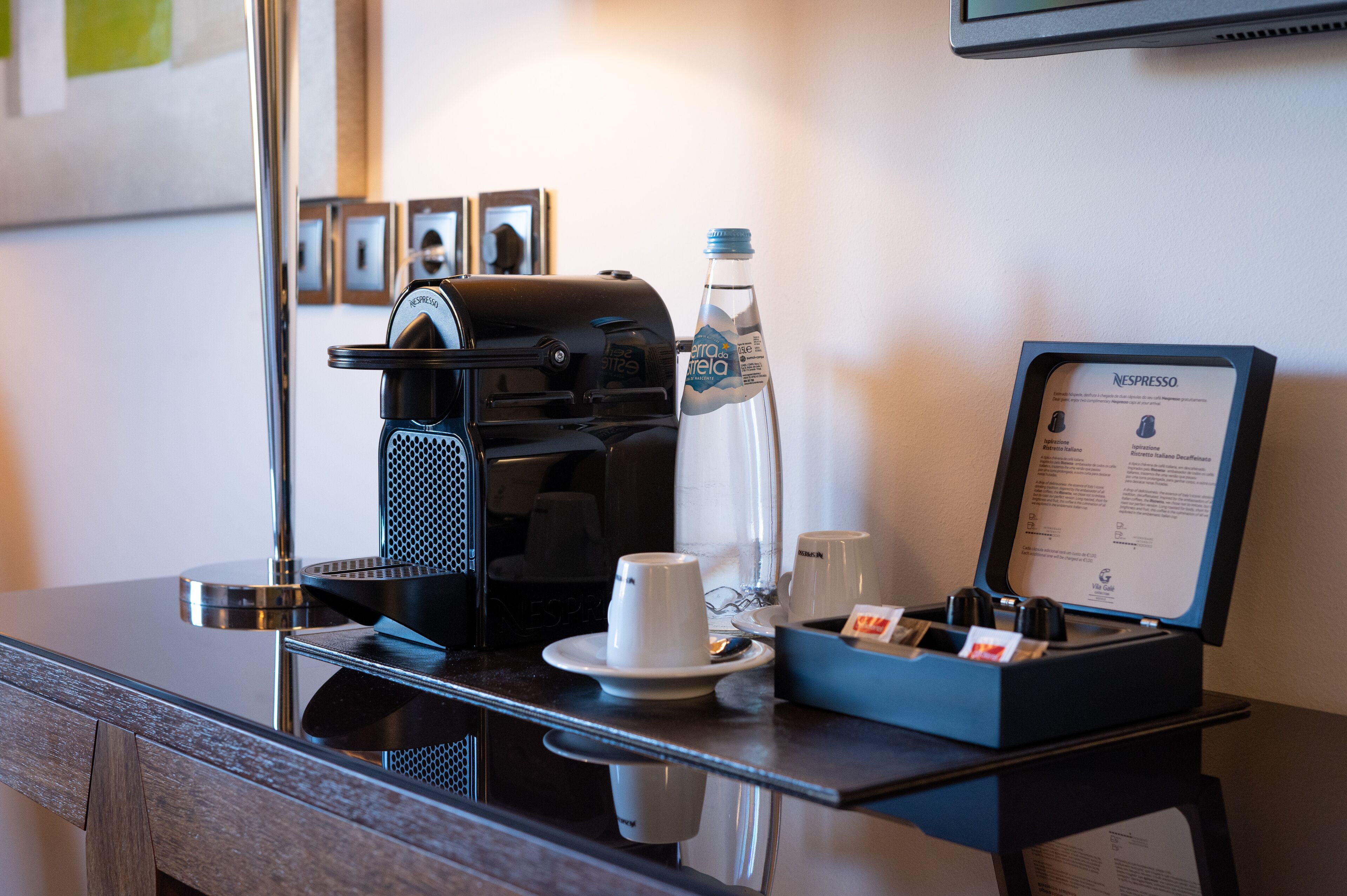 Estación de café en una habitación de hotel, con cafetera, botella de agua, tazas y un menú.