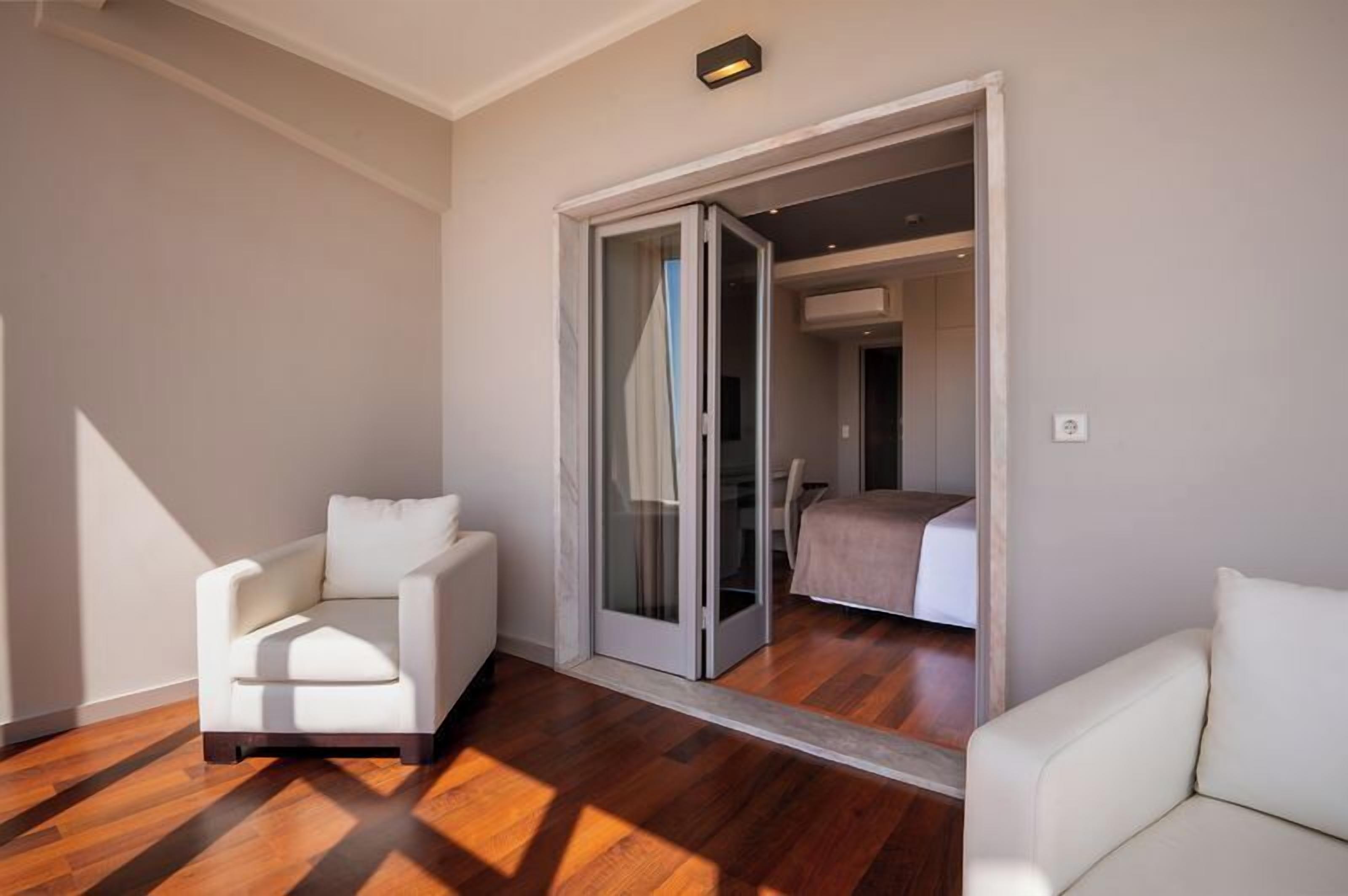 Una habitación con una silla blanca, un sofá blanco, una puerta corrediza de vidrio que da a un dormitorio y suelo de madera.